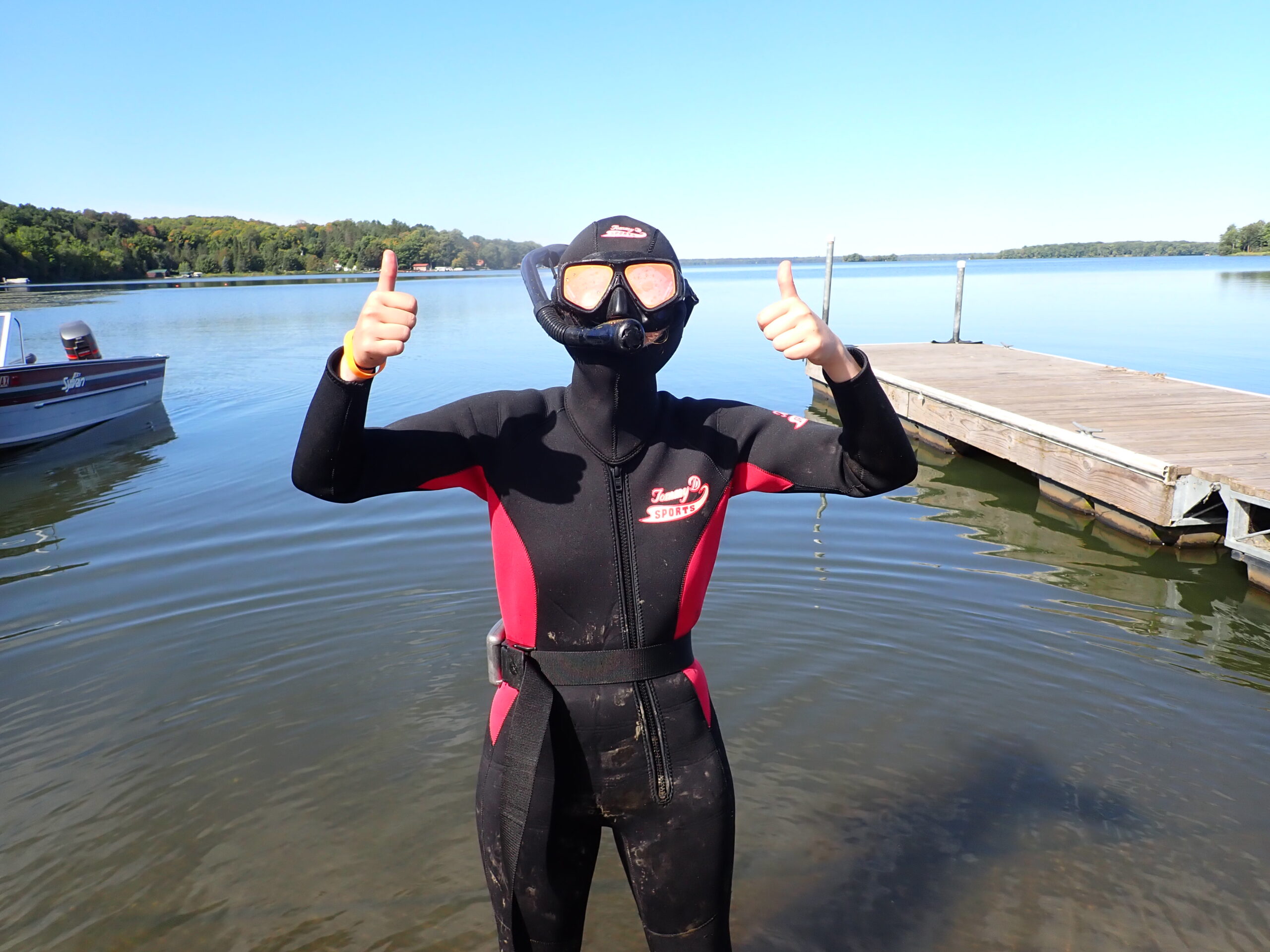 A figure in a full wetsuit with a hood and goggles stands in front of a lake giving two thumbs up. 