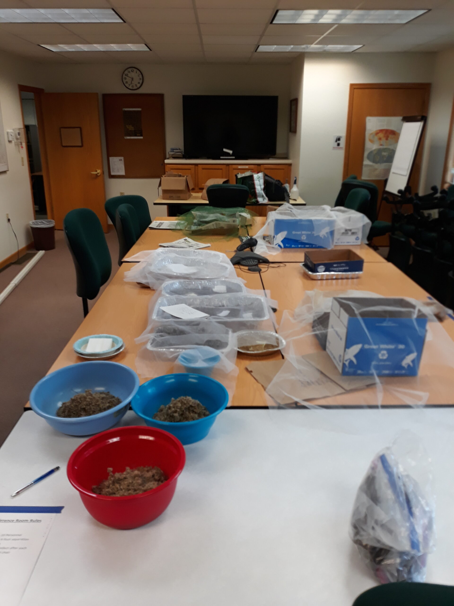 A large conference table is full of bowls, boxes, and pans. Each container holds native seed and most are covered with netting.